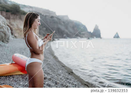 Woman, beach, phone. Smiling woman using smartphone on pebble beach during summer vacation with copy space. Woman, beach, phone. Smiling woman using smartphone on pebble beach during summer vacation with copy space. 139832353