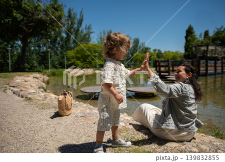Mother and son high-five by a pond in a sunny kids park sharing a happy family moment Mother and son high-five by a pond in a sunny kids park sharing a happy family moment 139832855