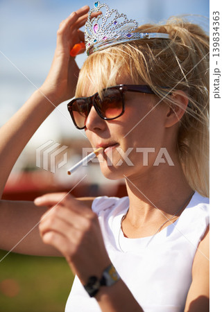 Crown, smoking and young woman with cigarette in nature at outdoor festival, event or party. Fun, tiara and female person with tobacco, sunglasses and casual fashion in outside park or field. 139834363