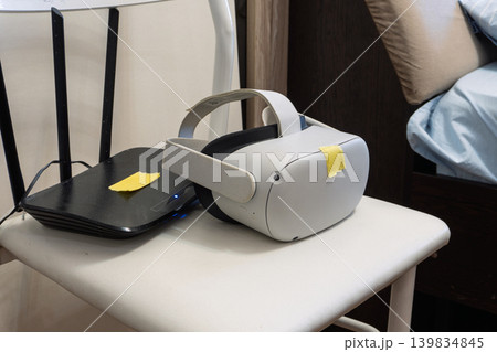 White virtual reality headset and black Wi-Fi router with yellow tape labels resting on a chair. Home network and gaming setup troubleshooting. Indoor technology equipment 139834845