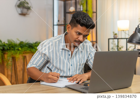 Indian man on remote learning uses laptop and notebook writes notes completes test quiz exam online Indian man on remote learning uses laptop and notebook writes notes completes test quiz exam online 139834870