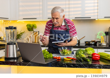 Senior old man making vegetable salad using laptop recipe and writing notebook notes for diet plan Senior old man making vegetable salad using laptop recipe and writing notebook notes for diet plan 139834979