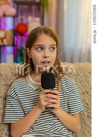 Child girl kid shyly sings alone into microphone sitting on sofa, smiling quietly with joy at home 139835068