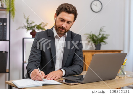 Middle-aged businessman attends webinar on laptop wears earphones takes notes snaps smartphone photo 139835121