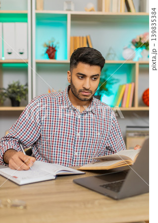Indian man on home table reading book and checking laptop while taking notes for online study lesson Indian man on home table reading book and checking laptop while taking notes for online study lesson 139835384