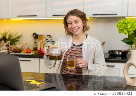 Young woman pouring hot tea in home kitchen enjoying cozy diet break with warm smile in calm ritual 139835421
