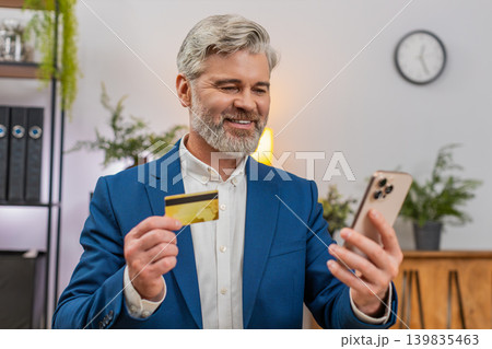 Middle-aged businessman confirms payment on smartphone holding credit card smiling at smooth service Middle-aged businessman confirms payment on smartphone holding credit card smiling at smooth service 139835463
