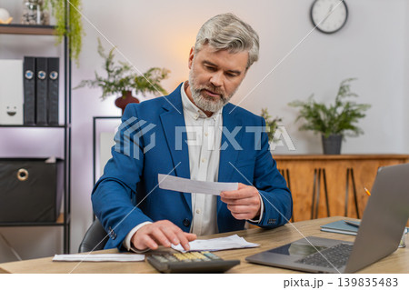 Middle-aged businessman reviews budget with laptop calculator receipts stack frowning over balance Middle-aged businessman reviews budget with laptop calculator receipts stack frowning over balance 139835483