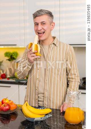 Middle-aged man eating banana in kitchen enjoying healthy eating for weight loss nutrition at home 139835495