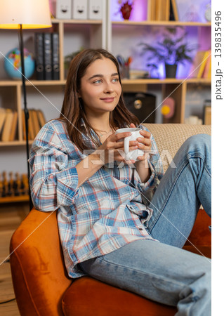 Young woman on home sofa enjoying hot drink and warming up in cozy living room during calm rest time Young woman on home sofa enjoying hot drink and warming up in cozy living room during calm rest time 139835496