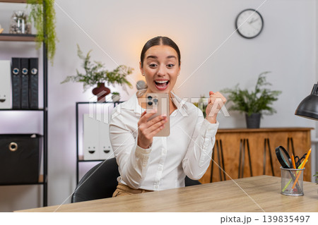 Young businesswoman typing fast on smartphone reads news of app game win and raises fist in victory Young businesswoman typing fast on smartphone reads news of app game win and raises fist in victory 139835497