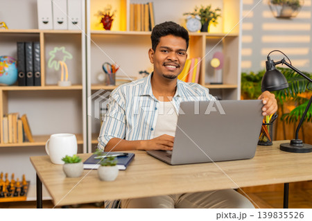 Indian man on home table opening laptop with cheerful smile and starting morning remote work indoors Indian man on home table opening laptop with cheerful smile and starting morning remote work indoors 139835526