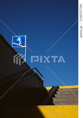 Urban scene featuring a blue underground pedestrian crossing sign against a clear deep blue sky. Concrete stairs with yellow edges lead upwards, creating a geometric composition with harsh shadows. Urban scene featuring a blue underground pedestrian crossing sign against a clear deep blue sky. Concrete stairs with yellow edges lead upwards, creating a geometric composition with harsh shadows. 139835794