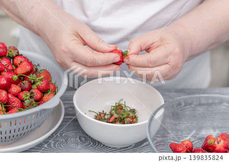 Women's hands brushing strawberries over a white bowl on a kitchen table 139835824