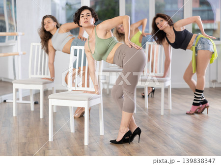 Energetic young female dancer practicing erotic movements of strip plastic near chair with other women in dance room 139838347