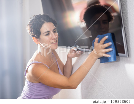 Positive young brunette woman cleaning dust from TV screen Positive young brunette woman cleaning dust from TV screen 139838605