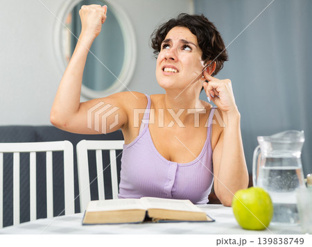 Displeased stressed woman suffering from noisy neighbors from above, covering his ears and shaking fist looking up while trying to reading book Displeased stressed woman suffering from noisy neighbors from above, covering his ears and shaking fist looking up while trying to reading book 139838749