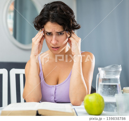 Young woman with headache sitting at table during reading book Young woman with headache sitting at table during reading book 139838786