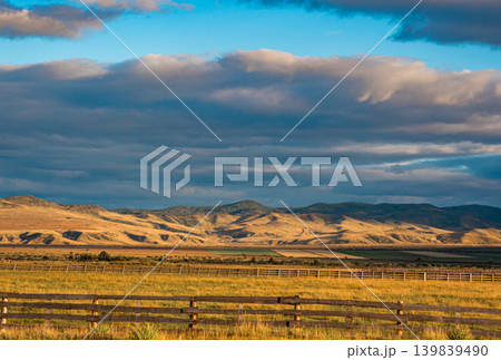 Expansive golden grasslands meet rolling hills under a dynamic sky. A long wooden fence crosses the foreground Expansive golden grasslands meet rolling hills under a dynamic sky. A long wooden fence crosses the foreground 139839490