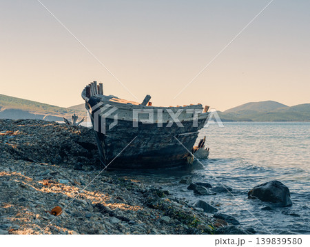 A decaying wooden boat, broken and weathered, lies abandoned on a stone shore A decaying wooden boat, broken and weathered, lies abandoned on a stone shore 139839580
