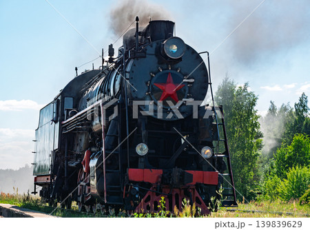 Black steam engine travels along railway track. The train passes through a vibrant green landscape with lush grass and a thick forest visible 139839629