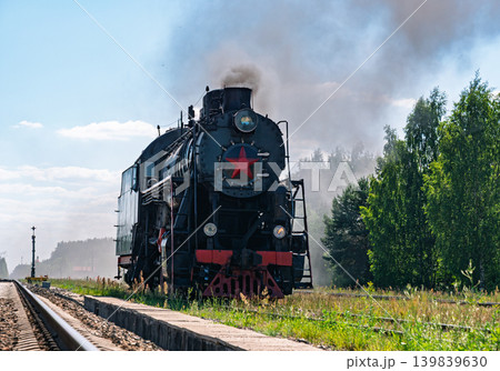 Black steam engine travels along railway track. The train passes through a vibrant green landscape with lush grass and a thick forest visible 139839630