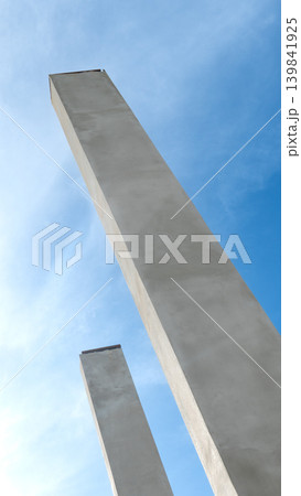 Low angle shot looking up at tall. Modern concrete poles or pillars stretching towards a bright blue sky with wispy white clouds. The structure highlights industrial construction. Low angle shot looking up at tall. Modern concrete poles or pillars stretching towards a bright blue sky with wispy white clouds. The structure highlights industrial construction. 139841925