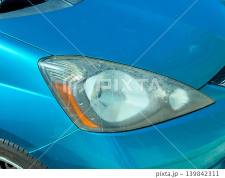 Headlight of a blue car showing signs of dirt and wear captured in bright light during the day near a parking area 139842311