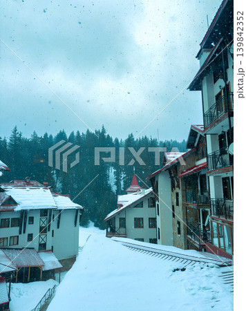 Snow falls on a quiet mountain resort as buildings stand under a cloudy sky with evergreen trees in the background during wintertime Snow falls on a quiet mountain resort as buildings stand under a cloudy sky with evergreen trees in the background during wintertime 139842352