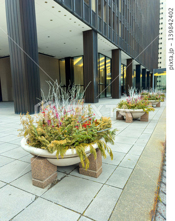 Planters with colorful plants sit on the sidewalk near a tall modern building in an urban area during a cloudy day 139842402