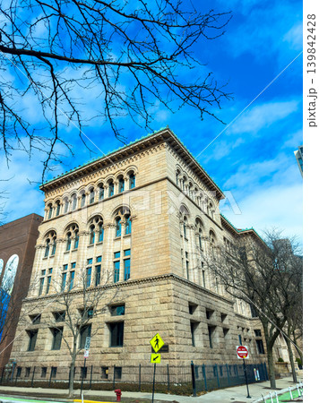 Historic stone building stands tall in urban setting under blue sky with light clouds and bare tree branches on a clear day Historic stone building stands tall in urban setting under blue sky with light clouds and bare tree branches on a clear day 139842428