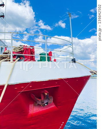 Red ship docked at the harbor with ropes, anchor, and green objects on deck under a blue sky with clouds 139842502