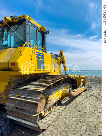 Bulldozer is parked on the beach during the day with a view of the ocean and ships in the background under a blue sky Bulldozer is parked on the beach during the day with a view of the ocean and ships in the background under a blue sky 139842548