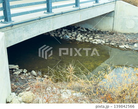 Bridge over shallow water with rocks and dry plants during daytime near an urban area in fall season Bridge over shallow water with rocks and dry plants during daytime near an urban area in fall season 139842566