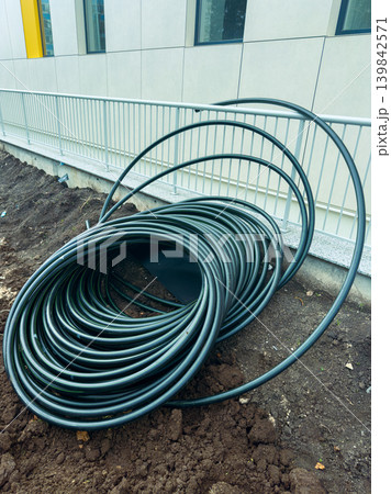 Several coils of black tubing are placed on the ground near a construction site while a gray building stands in the background under a cloudy sky Several coils of black tubing are placed on the ground near a construction site while a gray building stands in the background under a cloudy sky 139842571
