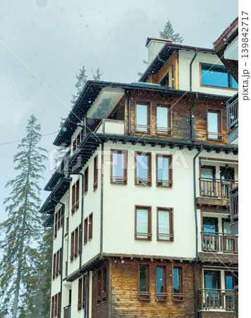 Snow falls on a mountain building in a chilly area surrounded by trees in the afternoon light during winter season 139842717