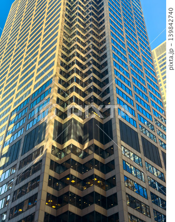Tall office building in a city with a repeating pattern on the facade showing reflections of the sky and surrounding structures during daytime hours 139842740