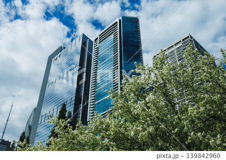 Modern buildings reflect clouds in the sky with trees in the foreground during a sunny day in a city Modern buildings reflect clouds in the sky with trees in the foreground during a sunny day in a city 139842960