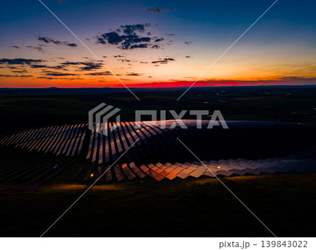 Sunset view of solar panels reflecting light on a clear evening near a rural area with open fields and distant hills Sunset view of solar panels reflecting light on a clear evening near a rural area with open fields and distant hills 139843022