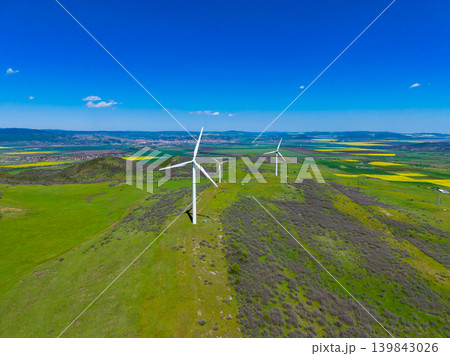 Wind turbines stand tall in a green field under a clear blue sky near a small town at midday in early spring Wind turbines stand tall in a green field under a clear blue sky near a small town at midday in early spring 139843026
