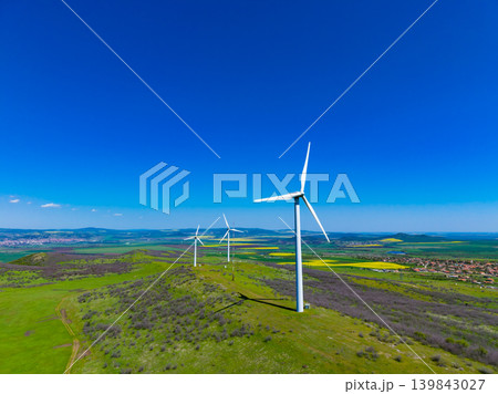 Wind turbines stand on a green hill under a blue sky in the countryside with fields and small buildings in the distance on a sunny day 139843027