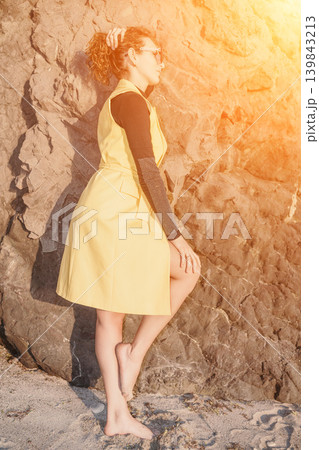 Woman yellow dress beach posing against a rocky cliff wall during a warm golden hour sunset with lens flare Woman yellow dress beach posing against a rocky cliff wall during a warm golden hour sunset with lens flare 139843213