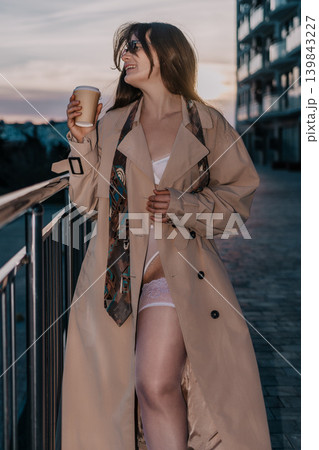 Woman coffee coat young lady holding paper cup wearing beige trench coat and lingerie on urban balcony at sunset Woman coffee coat young lady holding paper cup wearing beige trench coat and lingerie on urban balcony at sunset 139843227