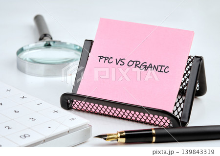 Pink note showing PPC versus Organic comparison rests in a holder on a white desk, alongside a calculator, magnifying glass, and pen for analysis 139843319