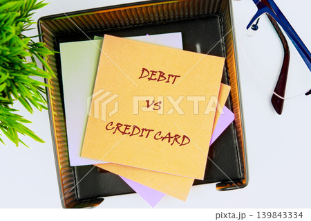 A yellow sticky note labeled Debit versus Credit Card sits atop other notes in a black tray on a white desk, next to a green plant and eyeglasses A yellow sticky note labeled Debit versus Credit Card sits atop other notes in a black tray on a white desk, next to a green plant and eyeglasses 139843334