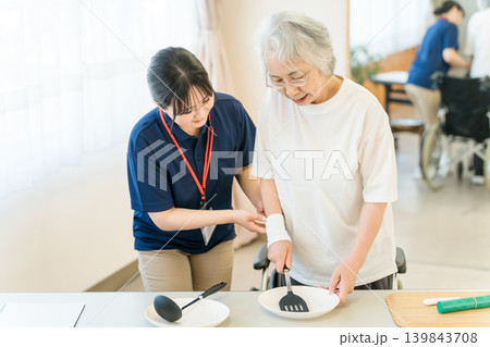 調理器具を使ってリハビリするシニア女性の補助をする作業療法士,ケアマネージャーの女性スタッフ 139843708