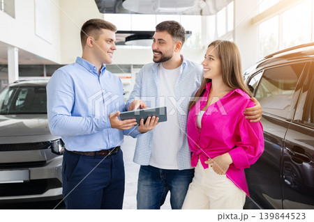 Professional salesman discussing automobile features with happy couple inside modern car dealership Professional salesman discussing automobile features with happy couple inside modern car dealership 139844523