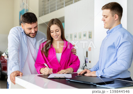 Smiling woman signing car purchase contract at dealership while partner embracing her Smiling woman signing car purchase contract at dealership while partner embracing her 139844546