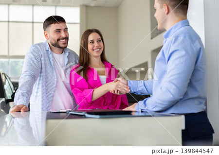 Smiling young couple shaking hands with car salesman at dealership desk Smiling young couple shaking hands with car salesman at dealership desk 139844547