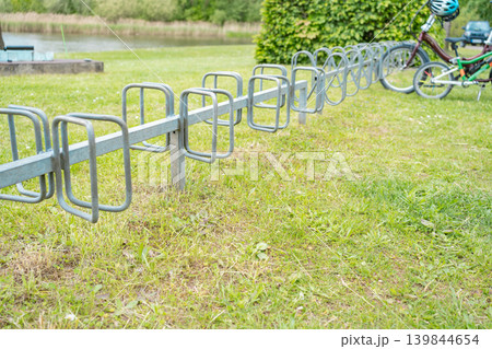 Steel bicycle stand structure on a green lawn near a recreation zone. Eco friendly transport support, urban design, sport infrastructure on a summer day. Steel bicycle stand structure on a green lawn near a recreation zone. Eco friendly transport support, urban design, sport infrastructure on a summer day. 139844654